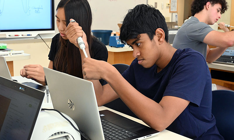 Two students at a lab desk with pipettes and laptop computers.