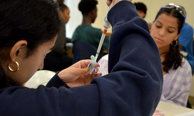Close view of student holding test tube and pipette with other students in the background.
